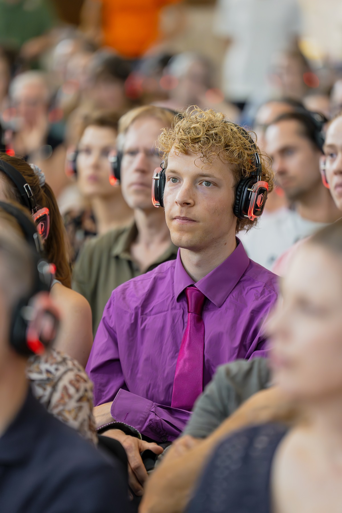 Een bezoeker met een paarse blouse en stropdas en met een koptelefoon op die aandachtig luistert naar een lezing.