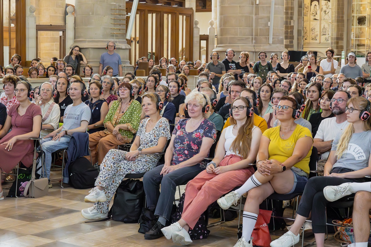 Publiek met koptelefoons op die geconcentreerd luisteren naar een lezing.